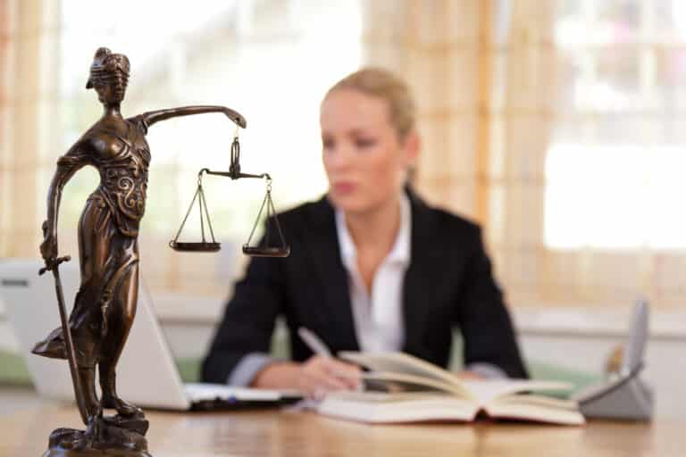 Scale of justice statue in foreground, female lawyer in background with laptop and book