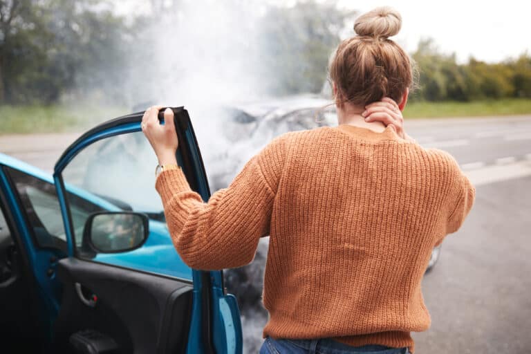 Young woman holding back of neck, standing beside car, staring at car accident wreckage