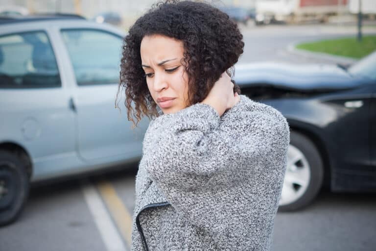 Woman holding the back of neck in front of car crash, indicating whiplash